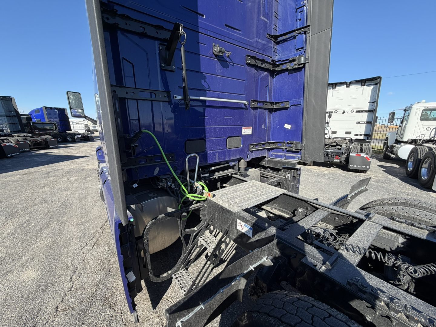 Truck cab and chassis area showing the back of a blue Mack PR64T76H sleeper with exposed frame and battery/air lines near the fifth wheel area.