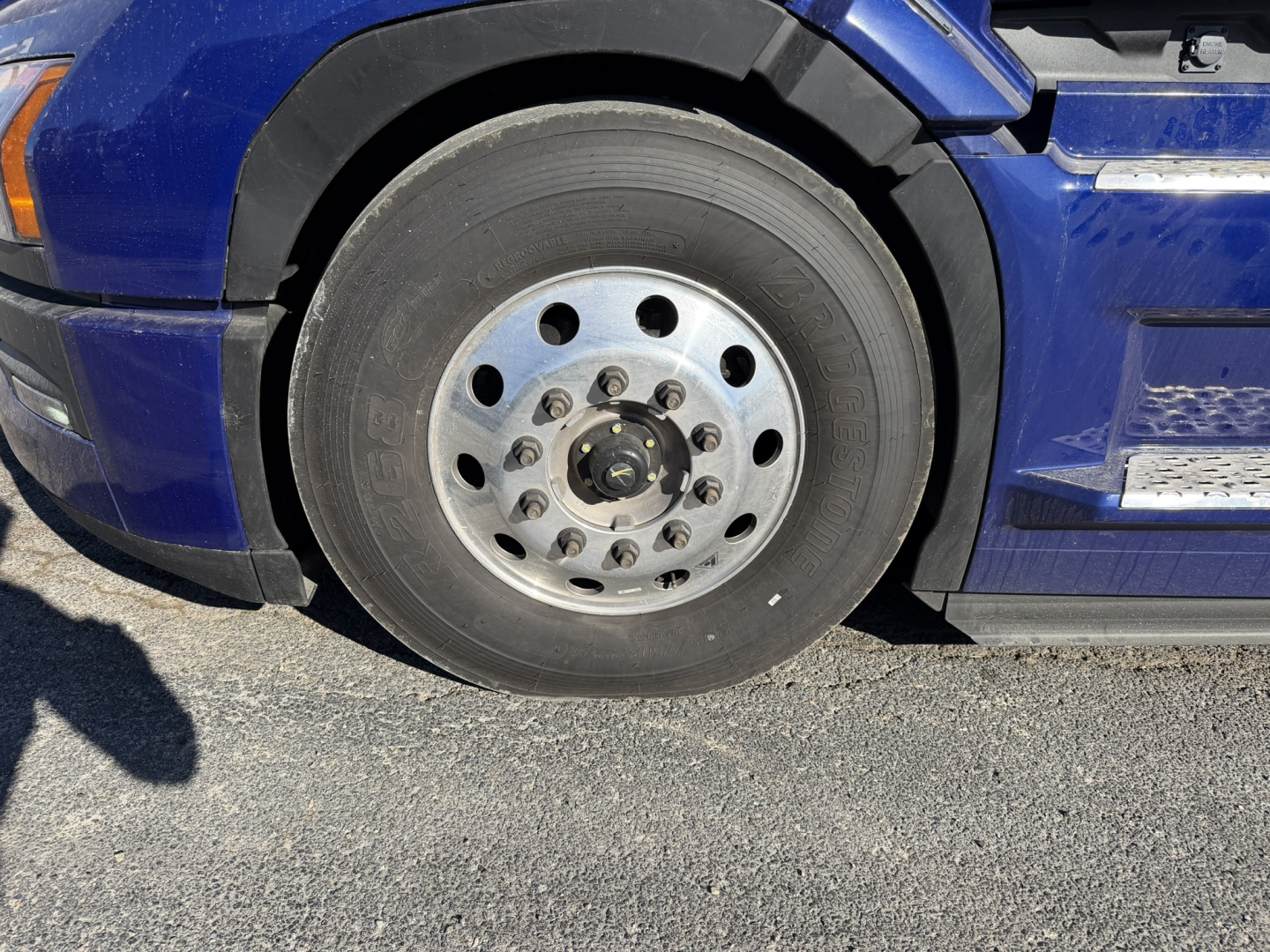Truck wheel and fender of a blue Mack PR64T76H showing a steel rim with multiple lug holes and a black tire.