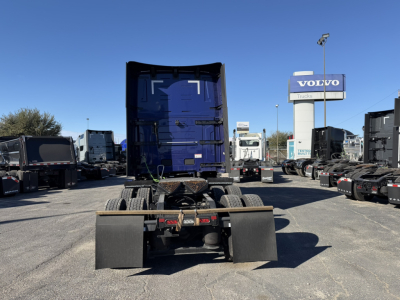Truck chassis rear view showing fifth-wheel and mounting hardware, blue cab in background, dealership lot with Volvo sign.