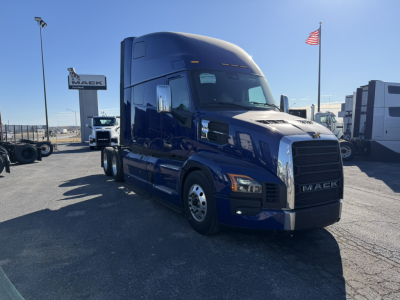 Truck cab side view of a blue 2026 Mack PR64T76H tractor in a dealership lot, front grille with MACK emblem, other trucks in background, American flag.