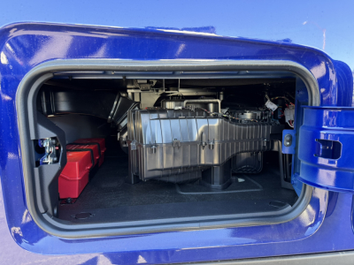Truck engine bay showing large chrome air intake, battery boxes, and surrounding blue body panel of a Mack PR64T76H conventional sleeper.