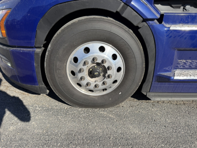 Truck wheel and fender of a blue Mack PR64T76H showing a steel rim with multiple lug holes and a black tire.