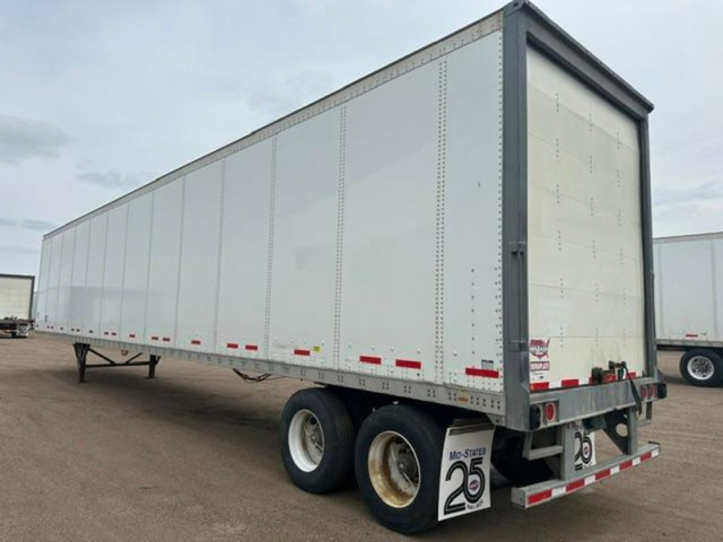 Trailer side view showing a white Wabash box trailer with dual rear axles and reflective red/white tape undercarriage.