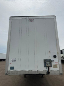 Rear view of a white 2016 Wabash trailer with riveted door, rear bumper, and electrical box; small decals and labels near bottom left; hitch/landing gear visible.