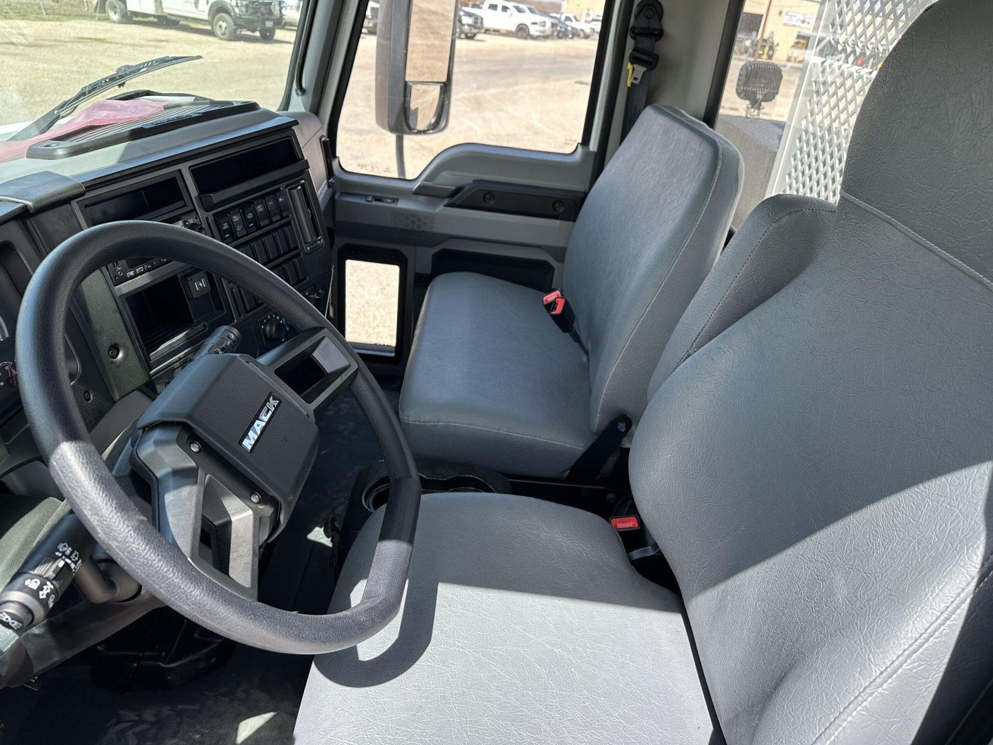 Seat and steering wheel area of a 2025 Mack MD7 cab, driver and passenger seats visible with gray upholstery; dashboard and windshield; side mirror reflection.
