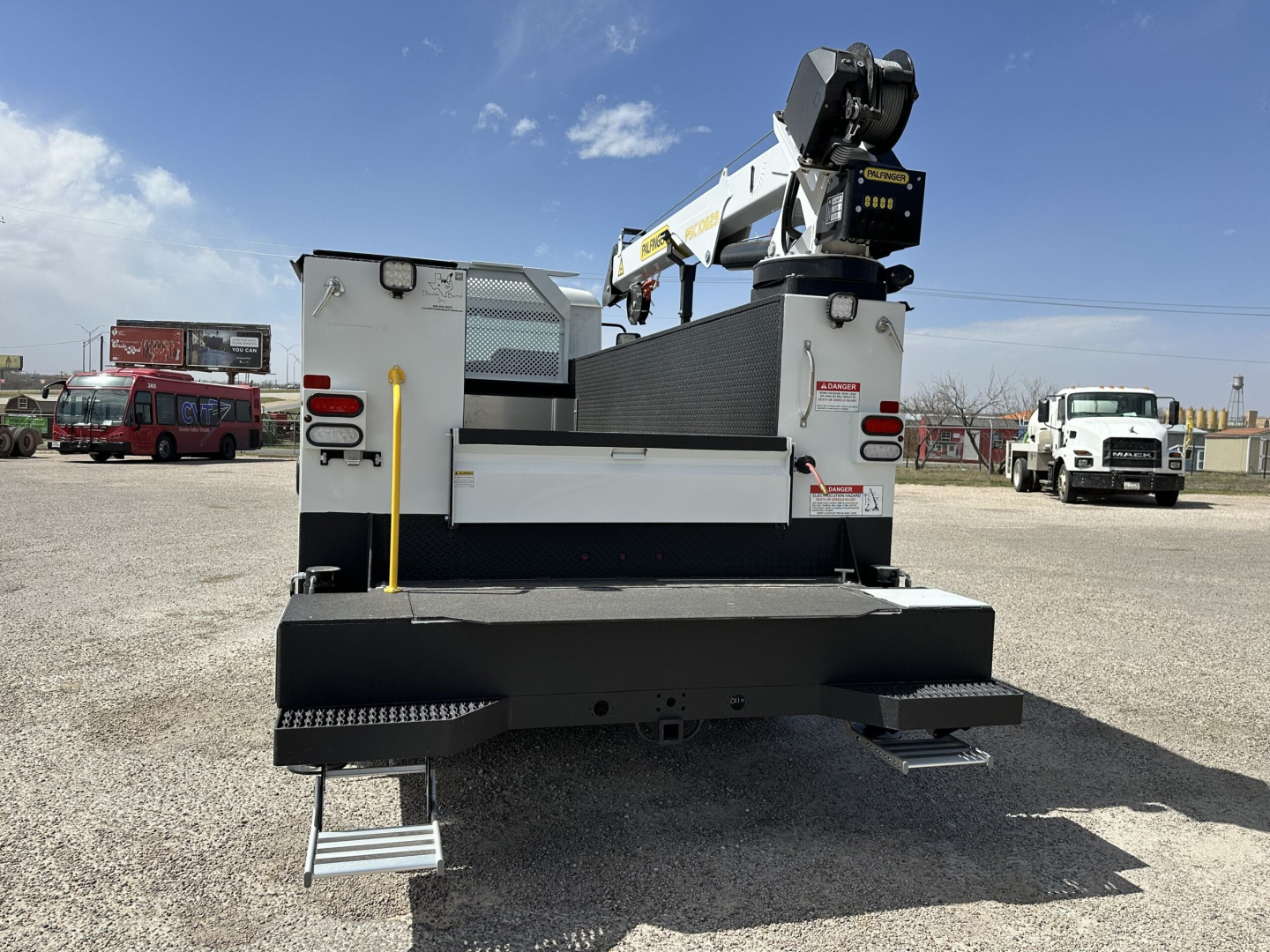 Rear view of a 2025 Mack MD7 service truck with mounted crane and hydraulic platform in a gravel lot.