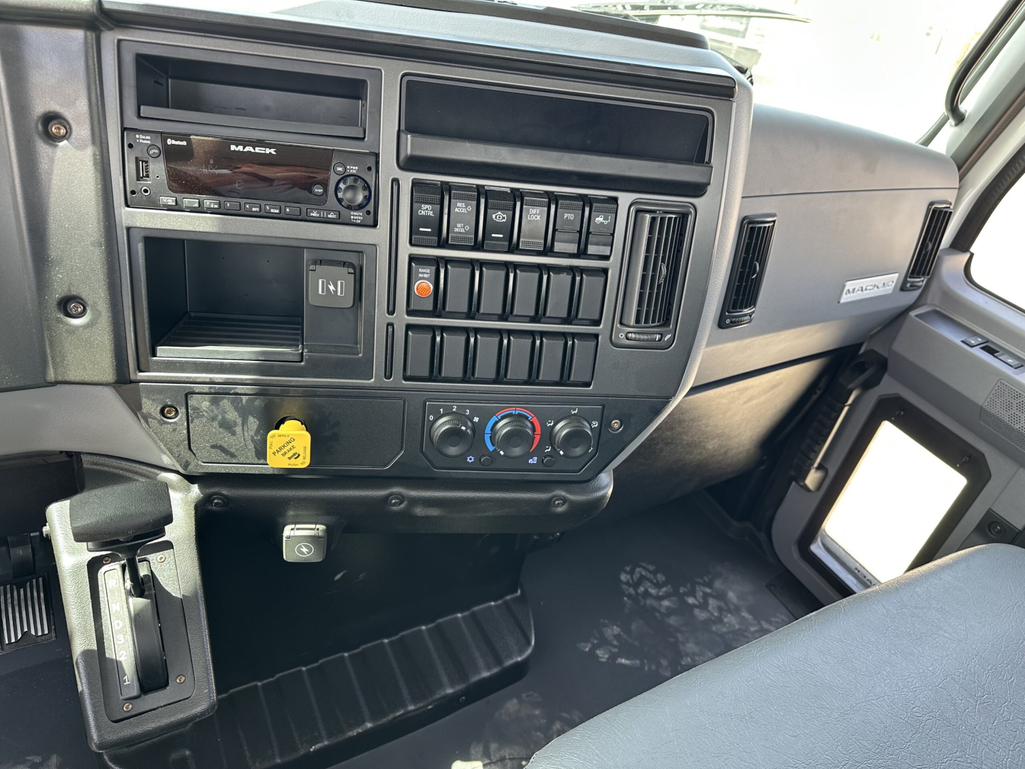 Cab interior of a Mack MD7 truck showing dashboard with stereo, multiple switches, climate controls, and a yellow knob in lower left.