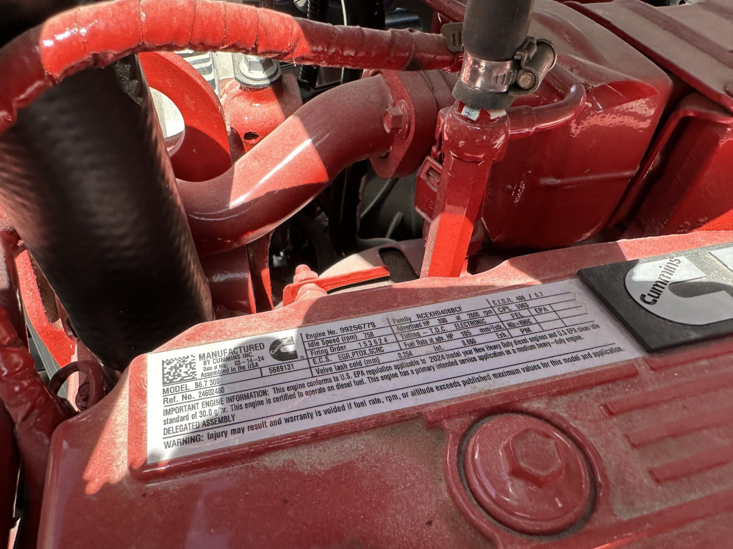 Engine bay of a 2025 Mack MD7 showing a label with specifications and fluid caps; red-painted components and hoses visible.