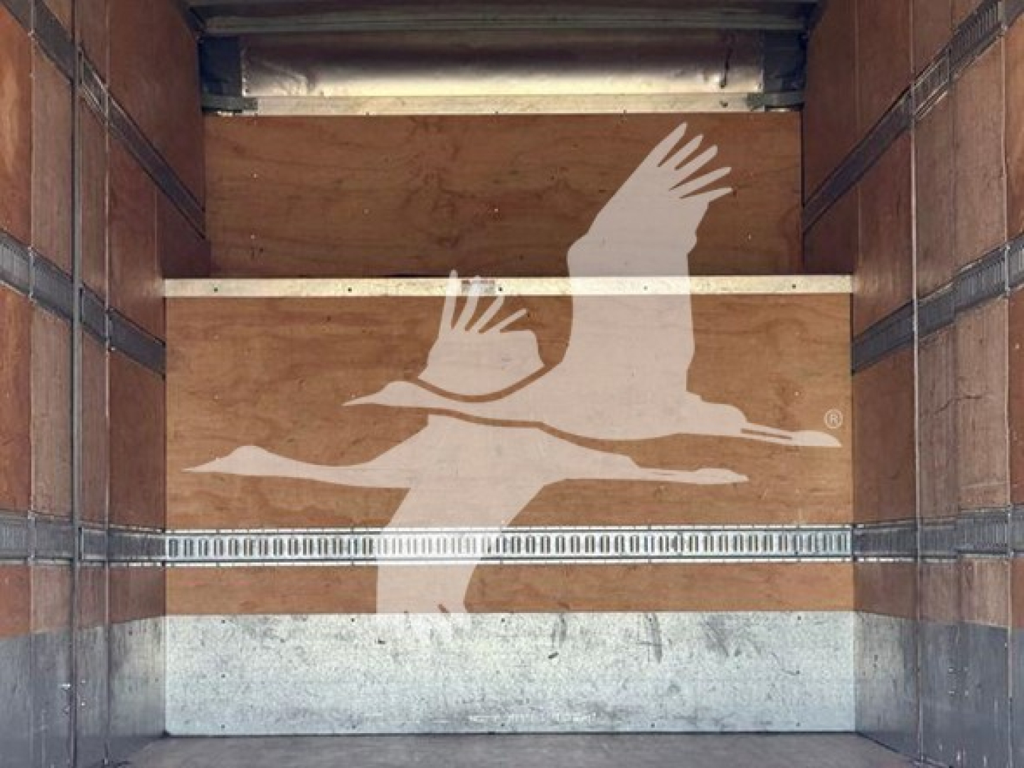 Trailer interior showing wooden walls with metal strap rails and a reflective logo of two flying geese on the side wall.