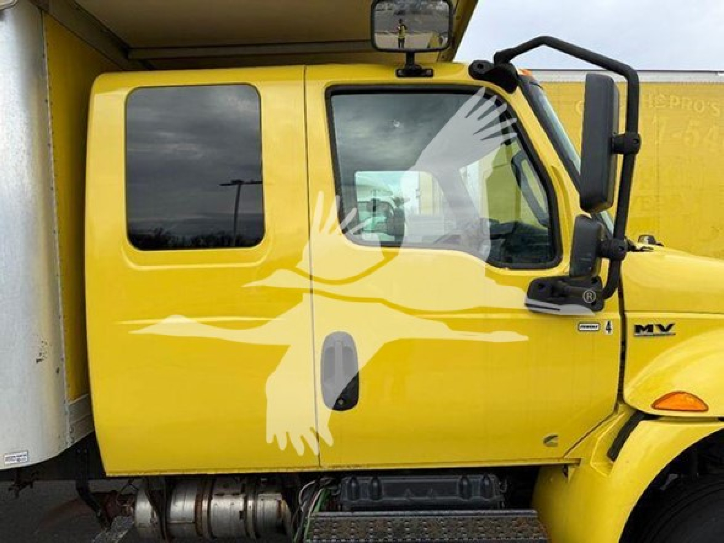 Yellow 2020 International MV moving van cab side with MV logo, window, mirror, and door handle visible.