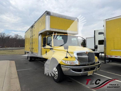 Truck in yard showing a yellow box truck body on a white forward cab with chrome bumper and grille. Side view reveals large cargo box and wheels; DOT/area lot in background.