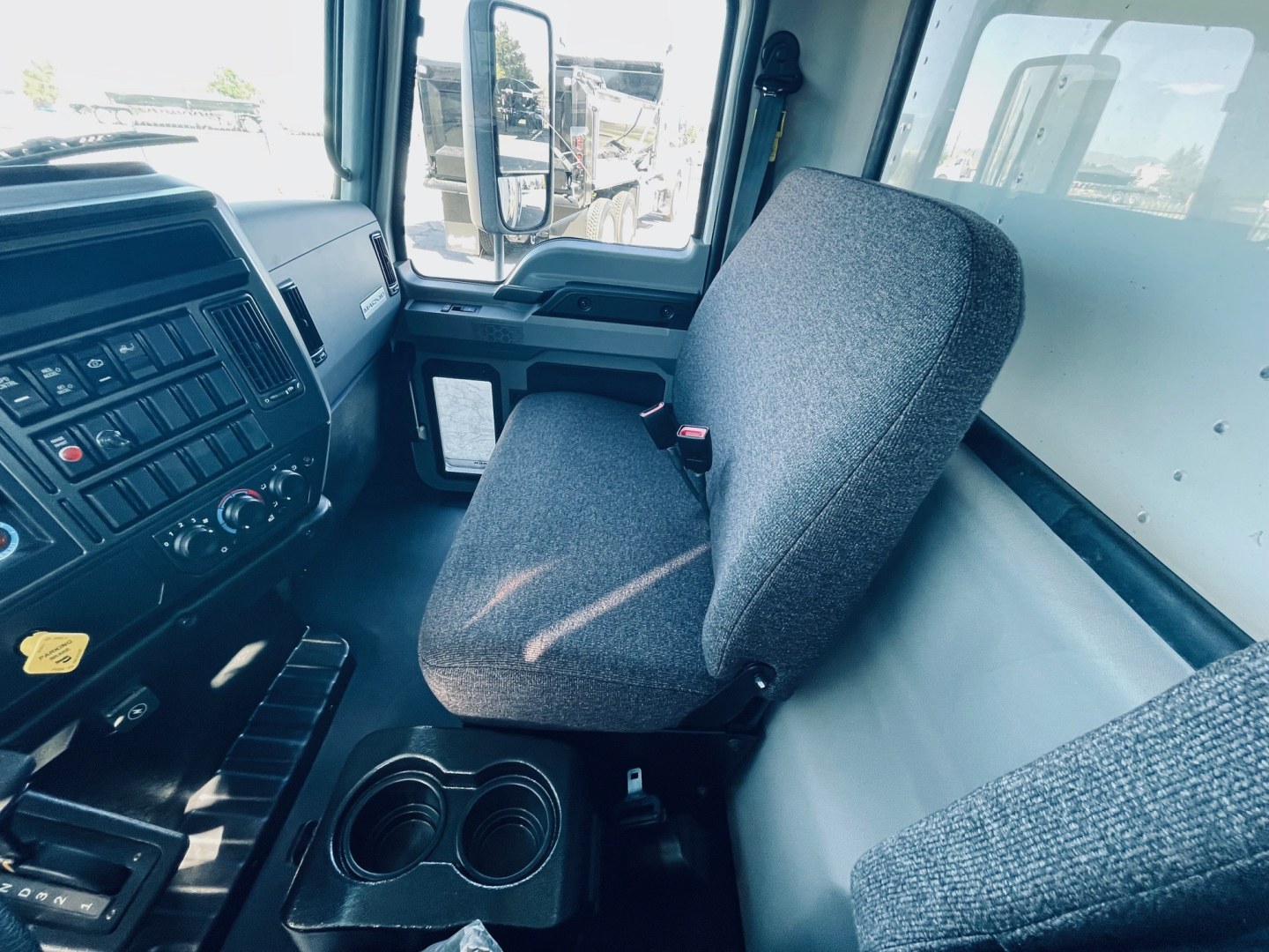 Truck cab interior showing driver seat and dashboard from left side, with controls and steering column visible; window view shows other trucks outside.