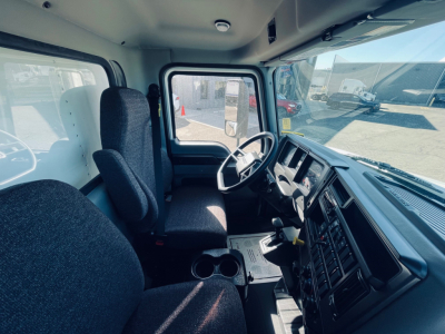 Truck cab interior of a 2026 Mack MD6 showing driver seat, steering wheel, dashboard, gear shifter, and center console with cupholders.