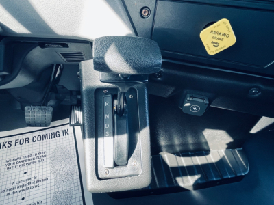 Dashboard area with gear selector showing R N D 3 and a parking brake yellow badge on dash; accelerator and brake pedals visible; some labels and paper mat in cabin.