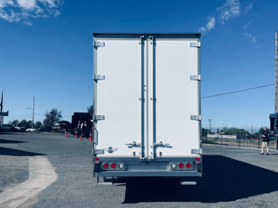 Rear view of a white 2026 Mack MD6 box truck with double rear doors, taillights, and bumper, in a parking lot.