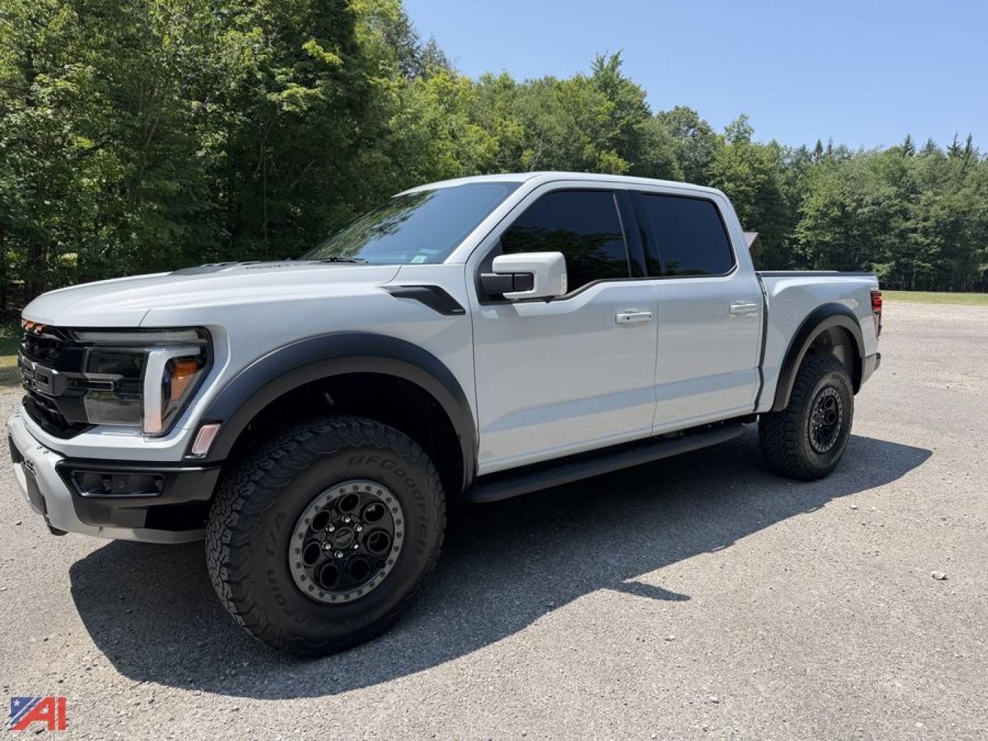 2025 Ford F-150 Raptor Crew Cab pickup truck in silver, angled three-quarter view. Large tires, black wheels, glossy body, running boards, 4x4 badge not visible. Clear sky and trees in background.