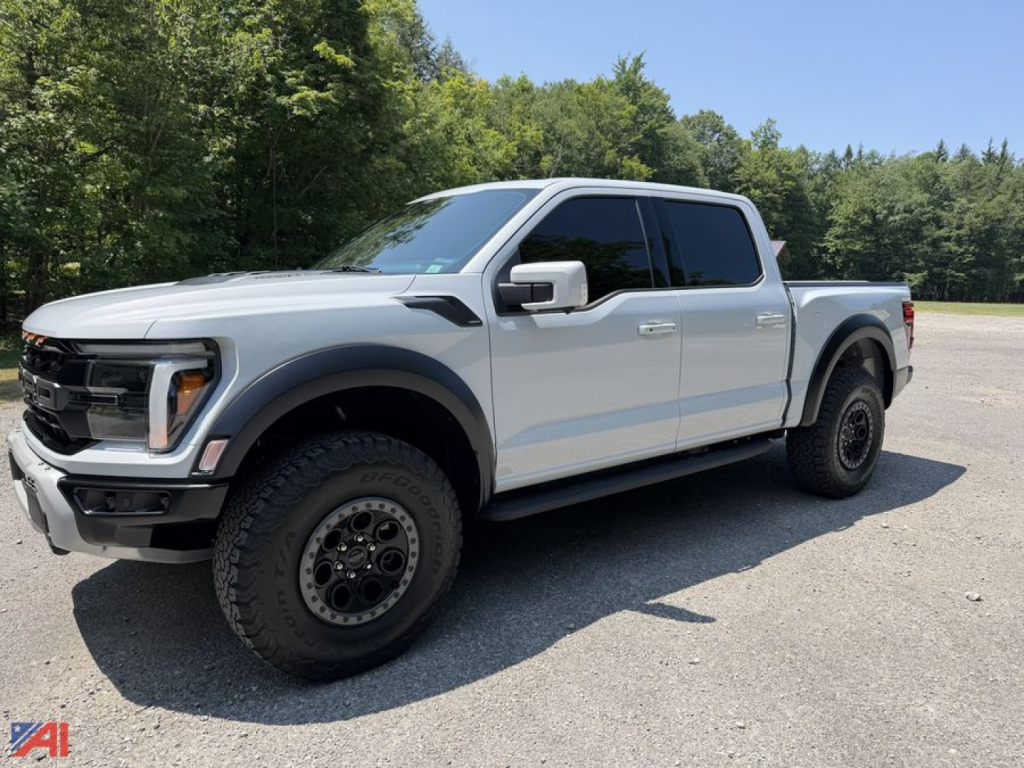 2025 Ford F-150 Raptor Crew Cab pickup truck in silver, angled three-quarter view. Large tires, black wheels, glossy body, running boards, 4x4 badge not visible. Clear sky and trees in background.