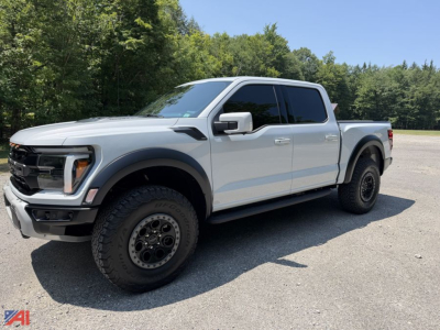 2025 Ford F-150 Raptor Crew Cab pickup truck in silver, angled three-quarter view. Large tires, black wheels, glossy body, running boards, 4x4 badge not visible. Clear sky and trees in background.