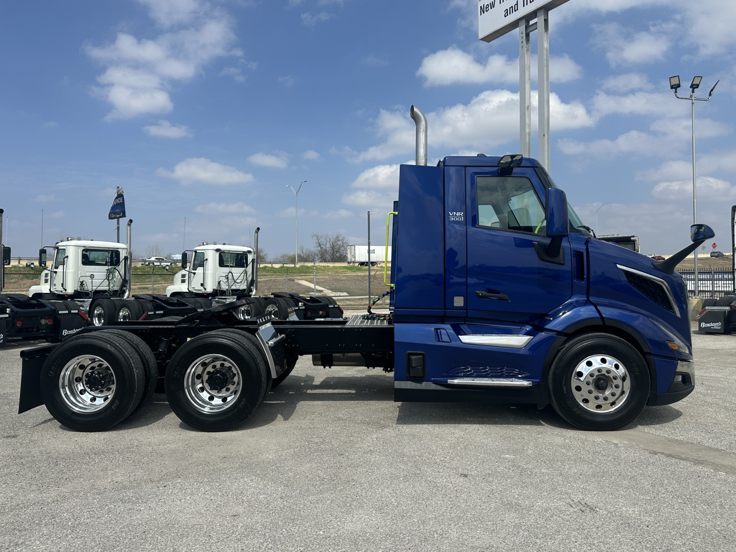 Volvo VNR64T300 tractor cab in blue viewed from side showing chassis and rear wheels, with two white trucks in background.