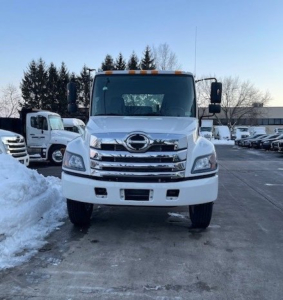 Front view of a white 2026 Hino L6 cab chassis truck showing chrome grille and bumper, in a dealership lot with snow piles.