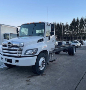 Truck cab chassis shows white Hino L6 cab with chrome grille and front bumper, flatbedless chassis behind, daylight setting, dealership lot in background.