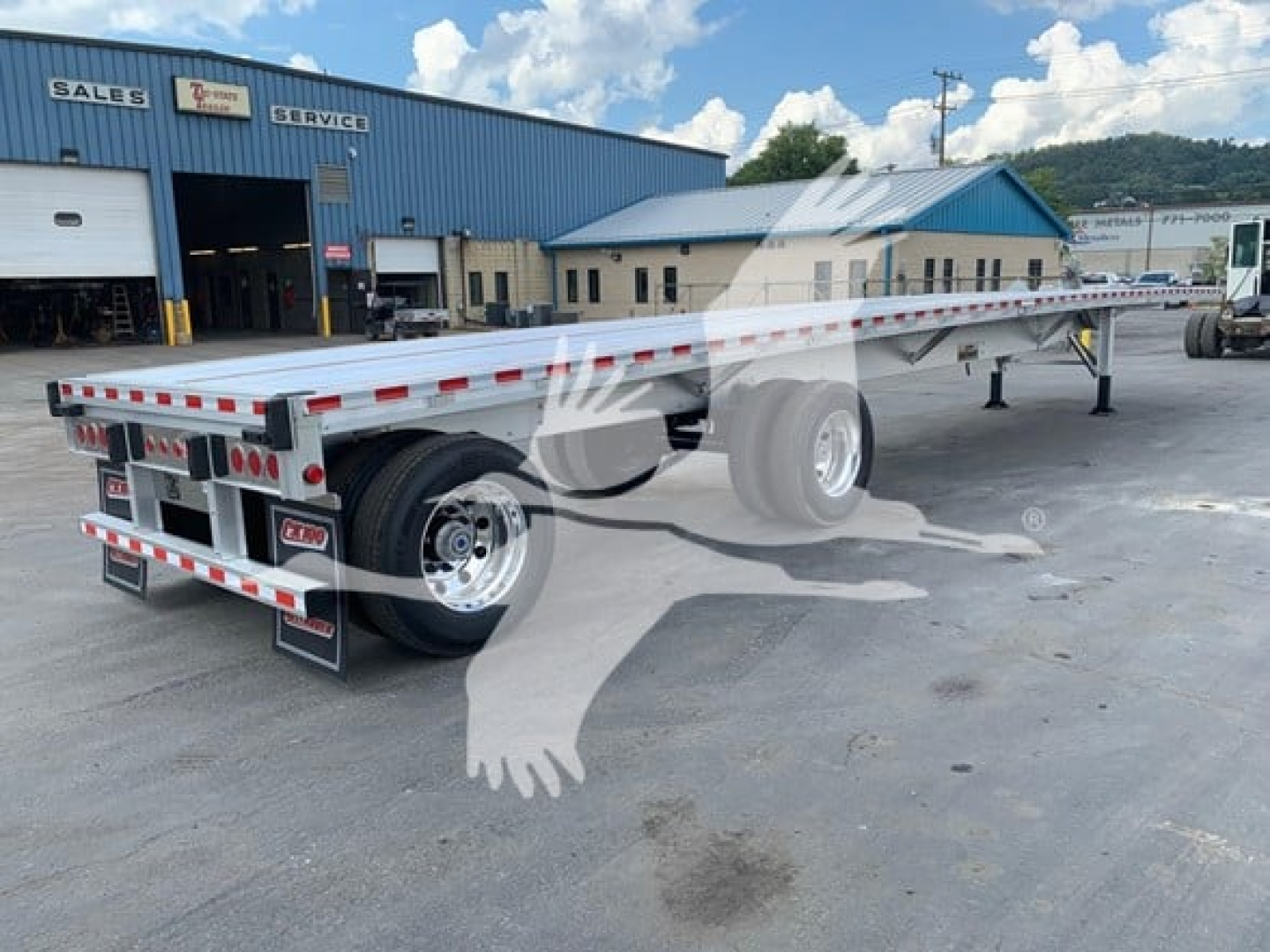 Flatbed trailer showing rear and side rails with dual wheels, red and white reflective tape, and a loading ramp area. Text on rear doors includes 'TR' and license plate area; background shows a blue service building.