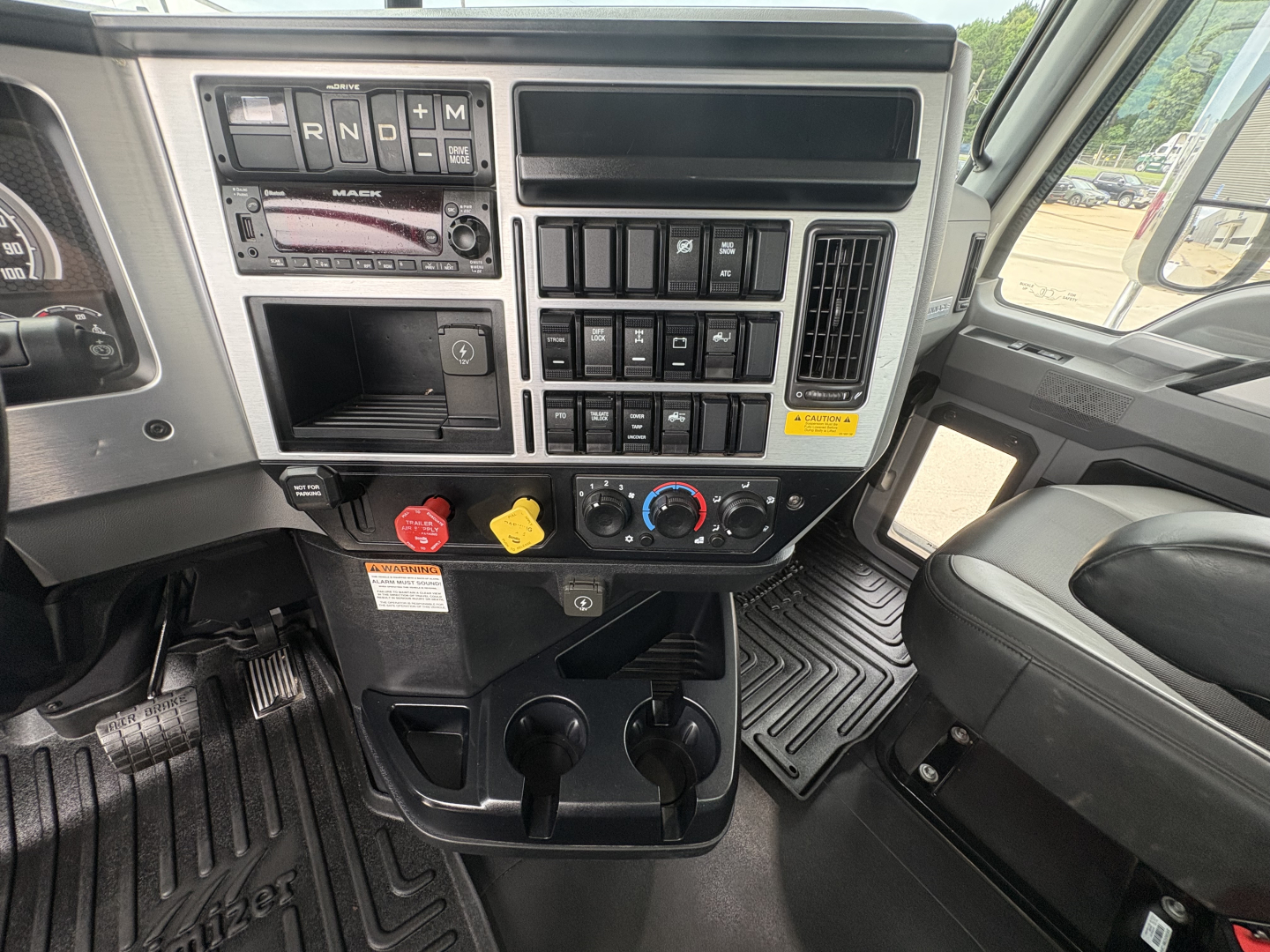 Mack truck cab interior showing dashboard controls, gear selector, warning switches, radio, climate controls, and cup holders.