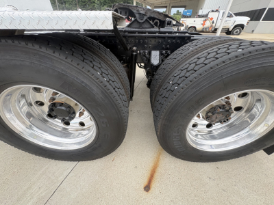 Twin rear axles of a Mack PI64T truck showing dual tires and wheel rims, with undercarriage components and rust stain on concrete.
