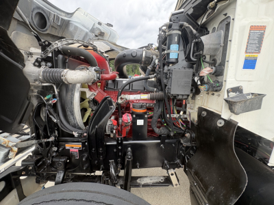 View into Mack PI64T engine bay showing diesel engine components, hoses, wiring, and chassis framing. Prominent red-painted components and black metal brackets visible.