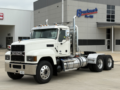 Truck front-left view of a white Mack PI64T daycab with chrome fuel tanks and stacked exhaust, parked in front of a showroom.