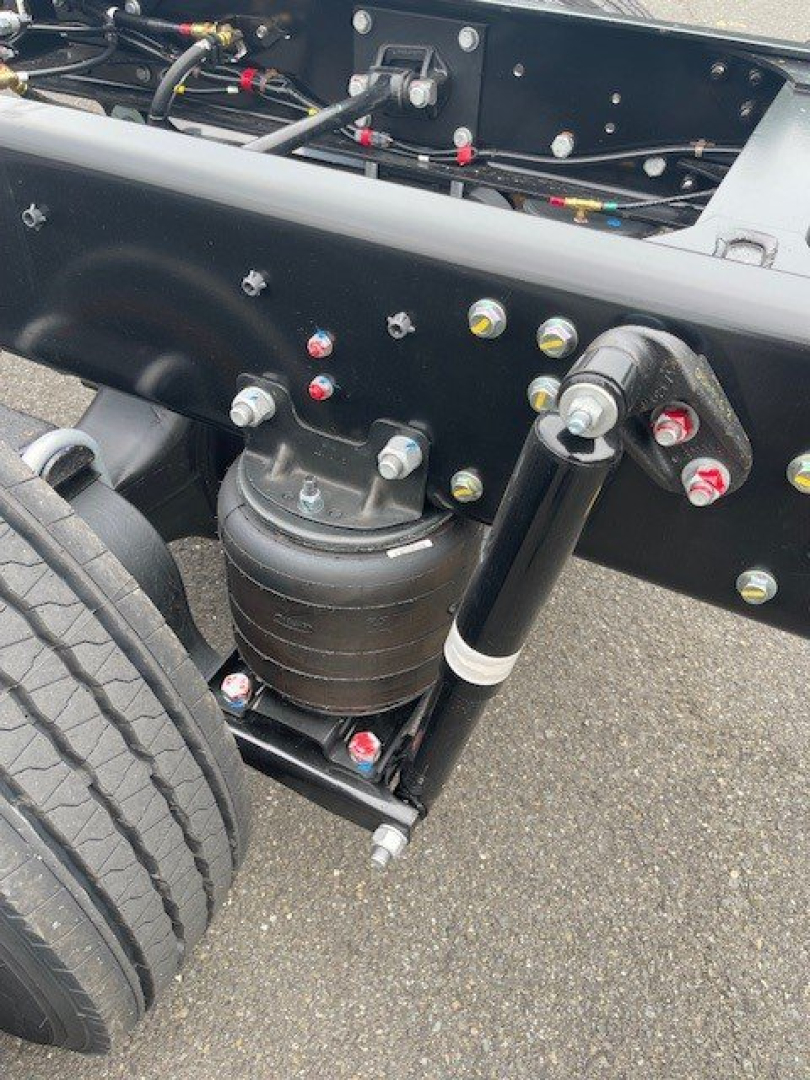 Truck front suspension and chassis showing air spring and hydraulic cylinder with bolts and hoses; visible components include a large black air spring, a shock/strut assembly, and various hydraulic lines on a black chassis plate.