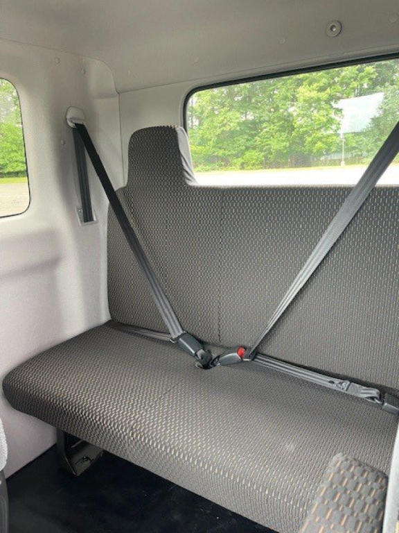 Truck cab interior rear seat with seat belts fastened, light gray upholstery, window view of trees; part of a 2027 Hino L6 cab chassis truck.