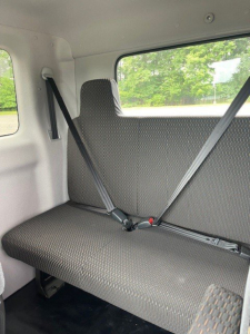 Truck cab interior rear seat with seat belts fastened, light gray upholstery, window view of trees; part of a 2027 Hino L6 cab chassis truck.