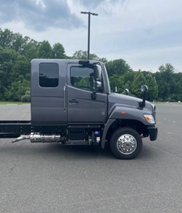 Cab chassis truck side profile showing gray Hino L6 with extended cab, dual rear wheels, and side fuel tank. Visible features include door, window, wheel, and mirror.