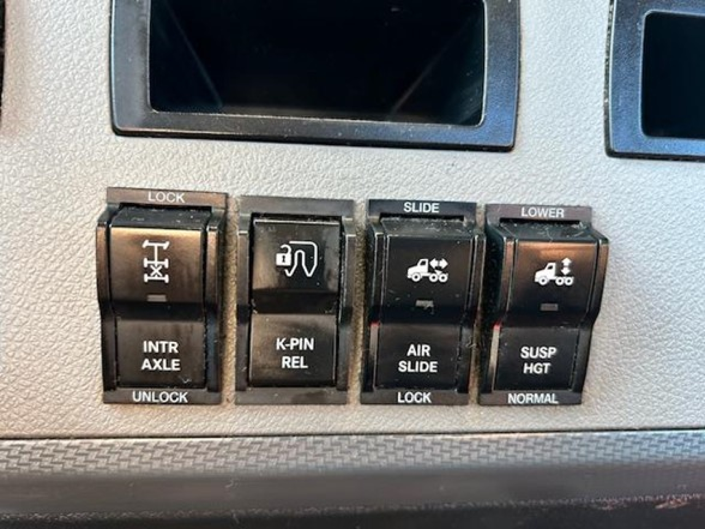 Dashboard switch panel showing lock,Intr Axle, K-PIN rel, air slide, lower suspension controls in a Freightliner Cascadia cabin.