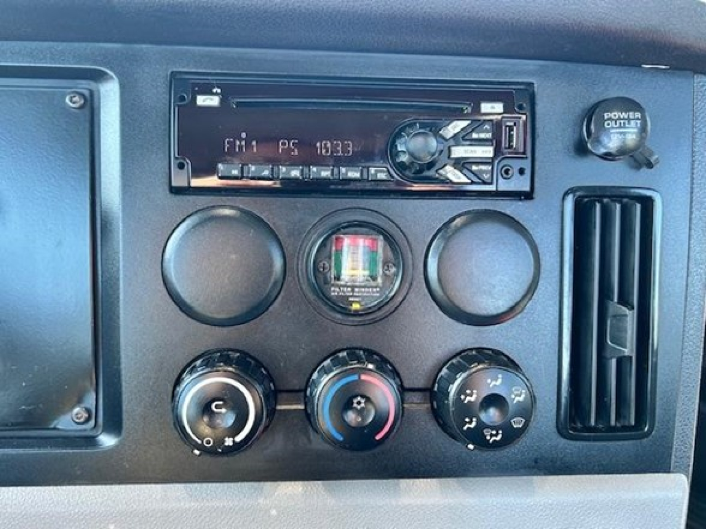 Truck dashboard panel showing climate/auxiliary controls and a single-DIN radio in a Freightliner Cascadia dash with round AC knobs and power outlet