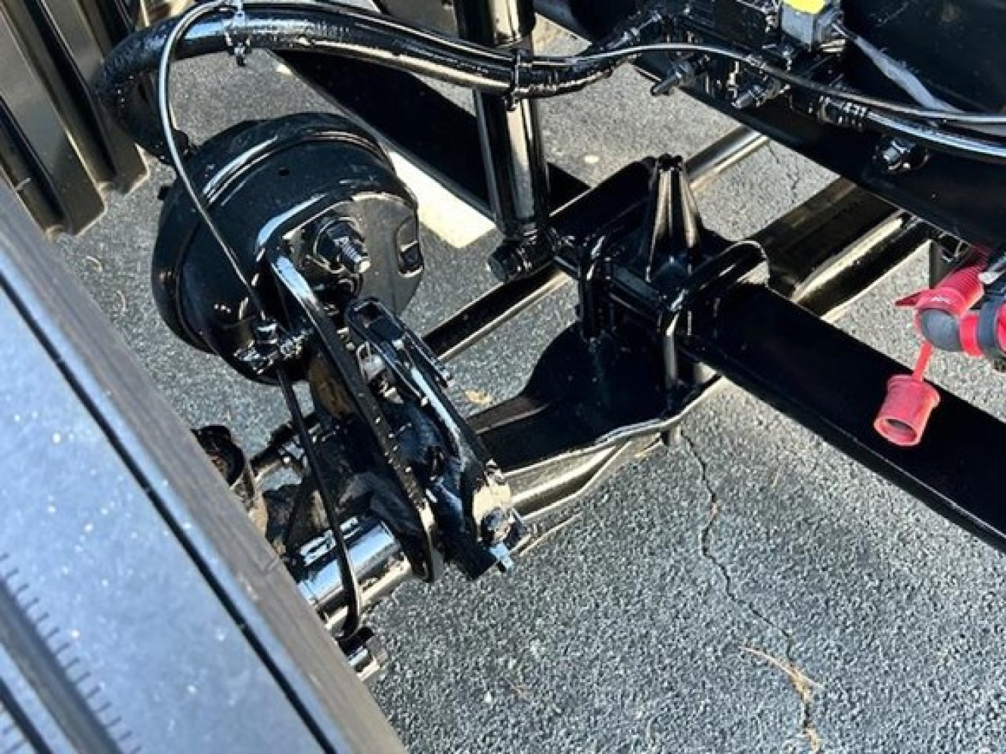 Rear undercarriage of a Freightliner Cascadia showing drivetrain components, with hoses and a black circular component, frame rails, and a red-capped fitting.