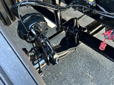 Rear undercarriage of a Freightliner Cascadia showing drivetrain components, with hoses and a black circular component, frame rails, and a red-capped fitting.