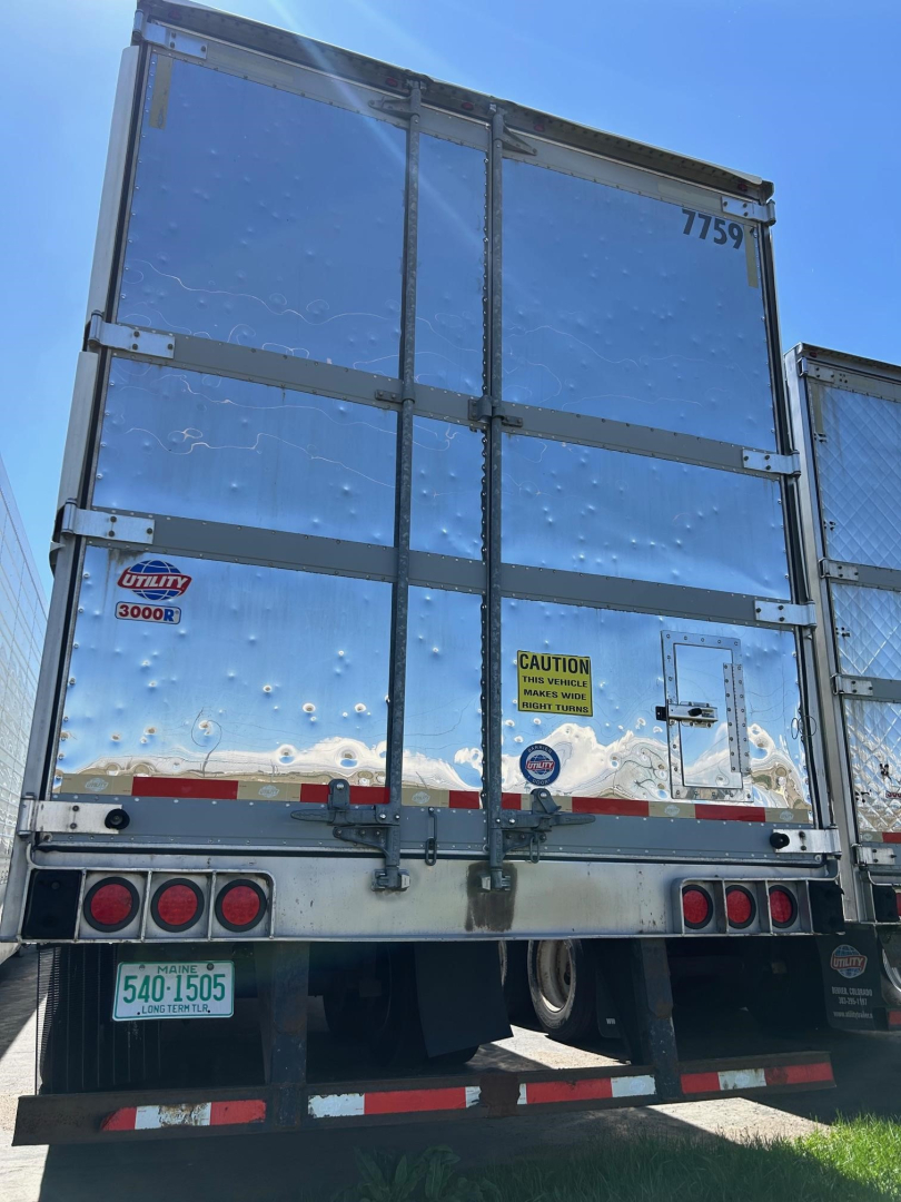 Rear view of 2018 Utility 3000R trailer showing reflective metal doors, bumper with red taillights, and stickers including 'Utility 3000R' and caution sign.