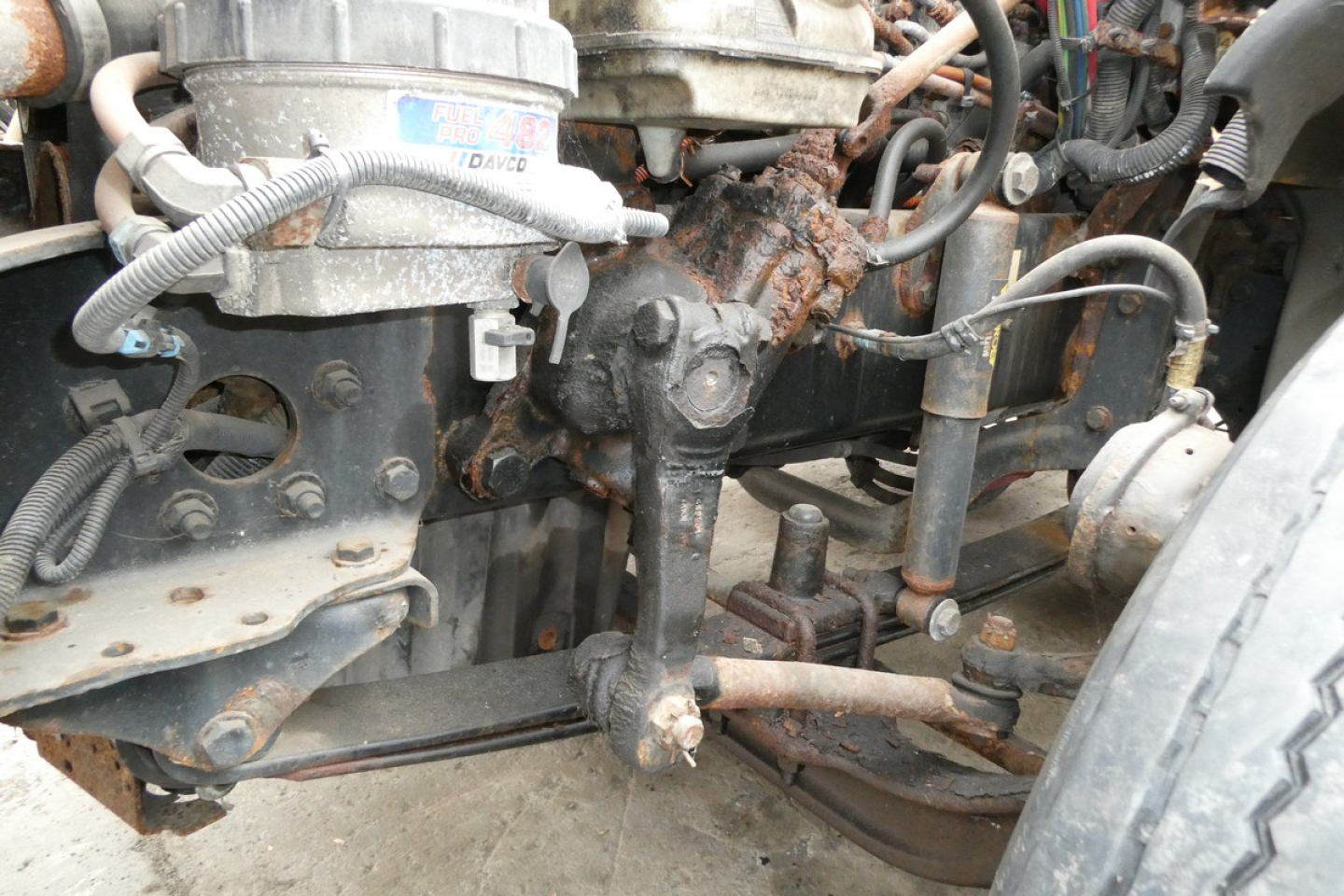Front left engine bay showing steering linkage and radiator hose with metal components and rusted mounting brackets.