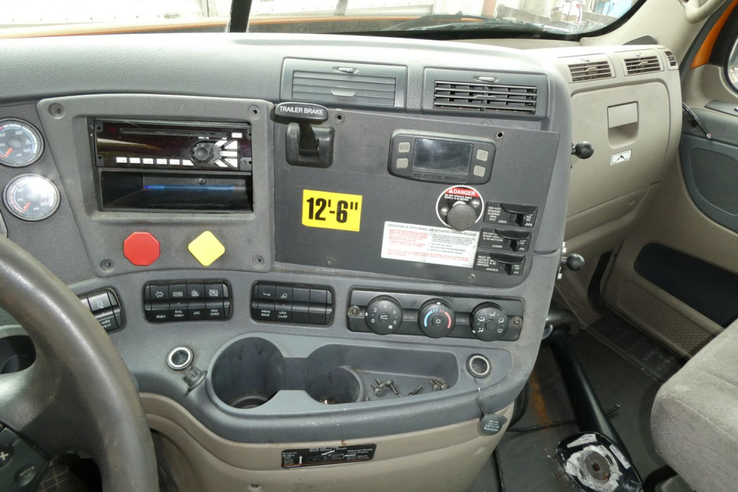 Dashboard of a Freightliner Cascadia 125 cab showing control cluster, radio slot, cup holders, and yellow 12-6" sticker.