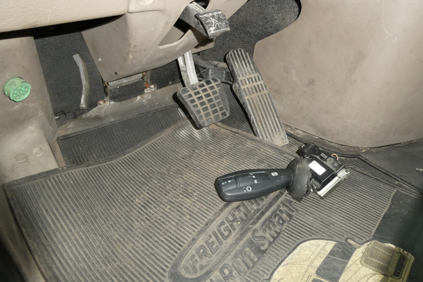Cab area of a Freightliner Cascadia 125 with pedals, steering column, and floor mat showing a key fob and switch.