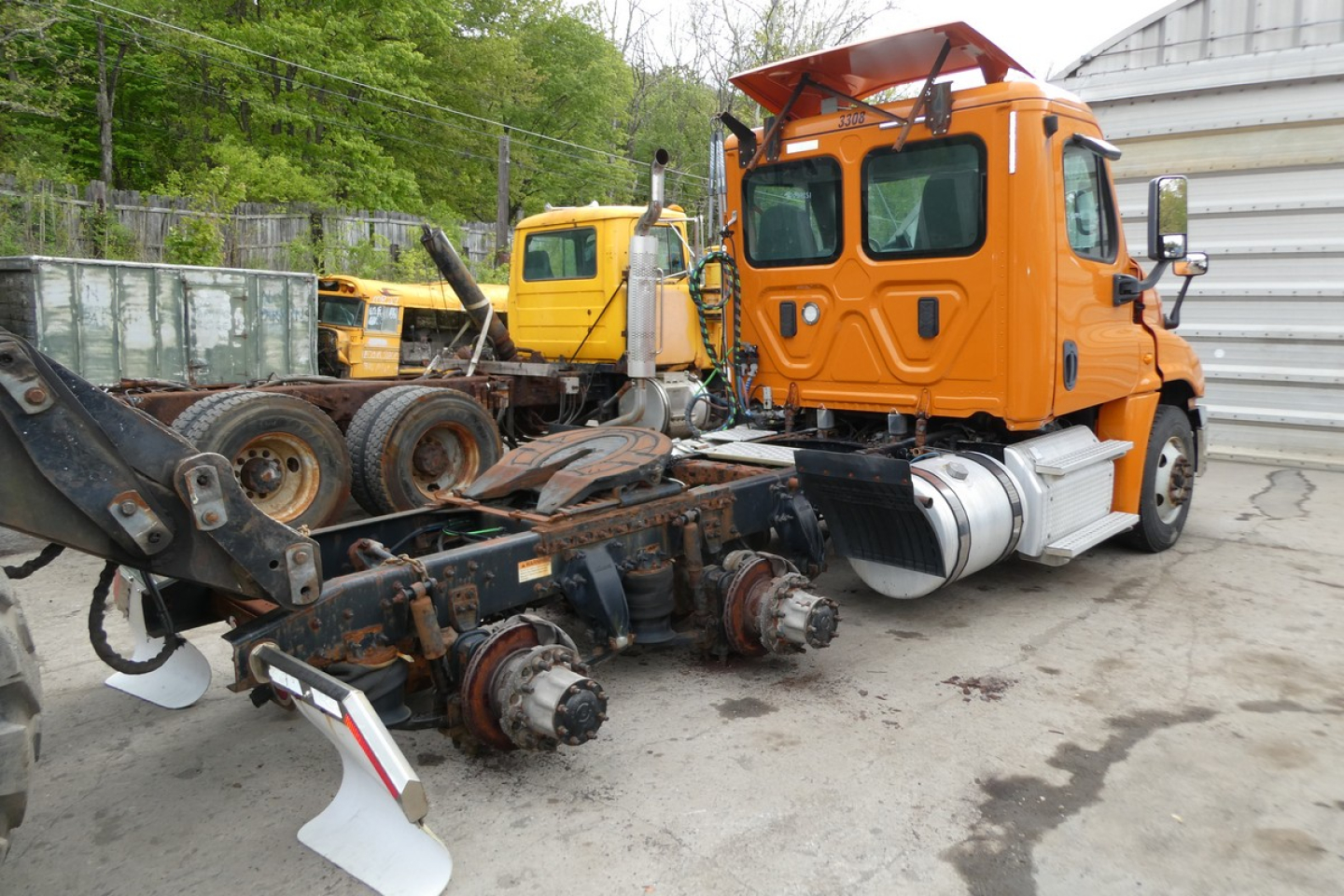 Orange Freightliner cab visible with attached chassis and wheel components laid on ground; other yellow trucks in background; no legible text on labels.