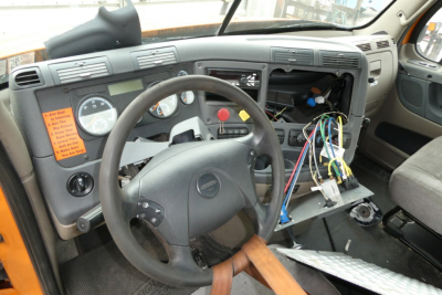 Cab interior of a Freightliner Cascadia daycab showing steering wheel, instrument cluster, exposed wiring, and dashboard with panels removed.