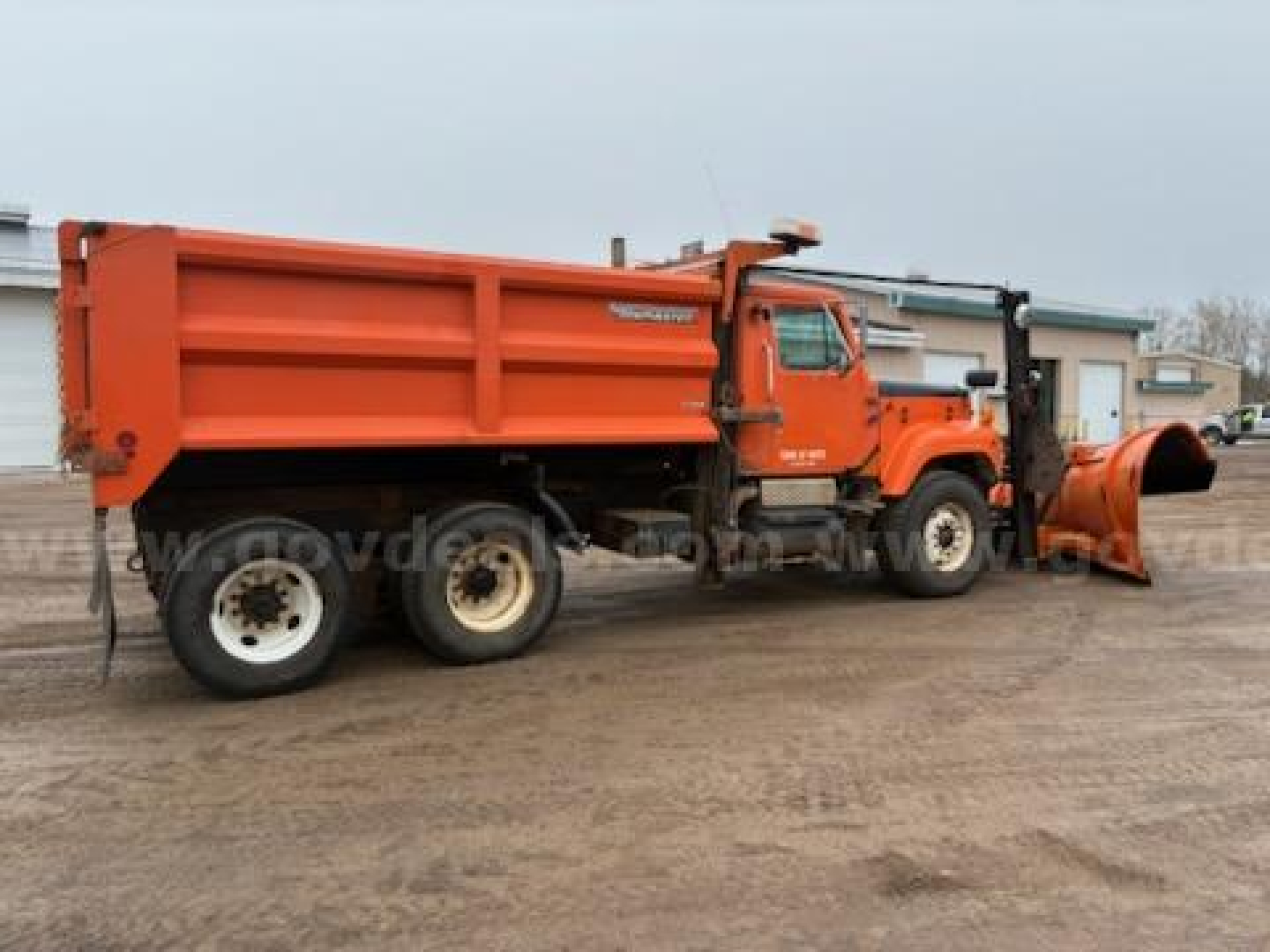 Big orange dump truck with plow attachment on front, International brand, dual rear wheels, side dump bed, headlights and cab visible.