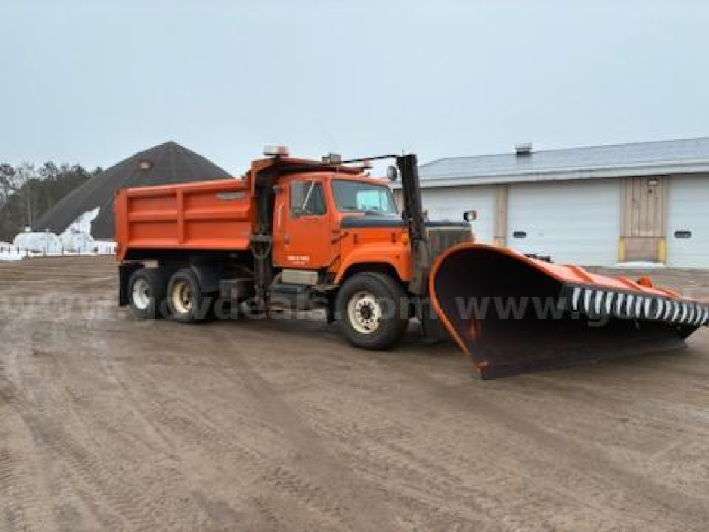Dump truck with a large orange snow plow/scoop attachment in front, International 2574 model visible, utility truck with hydraulic bucket and dump bed.