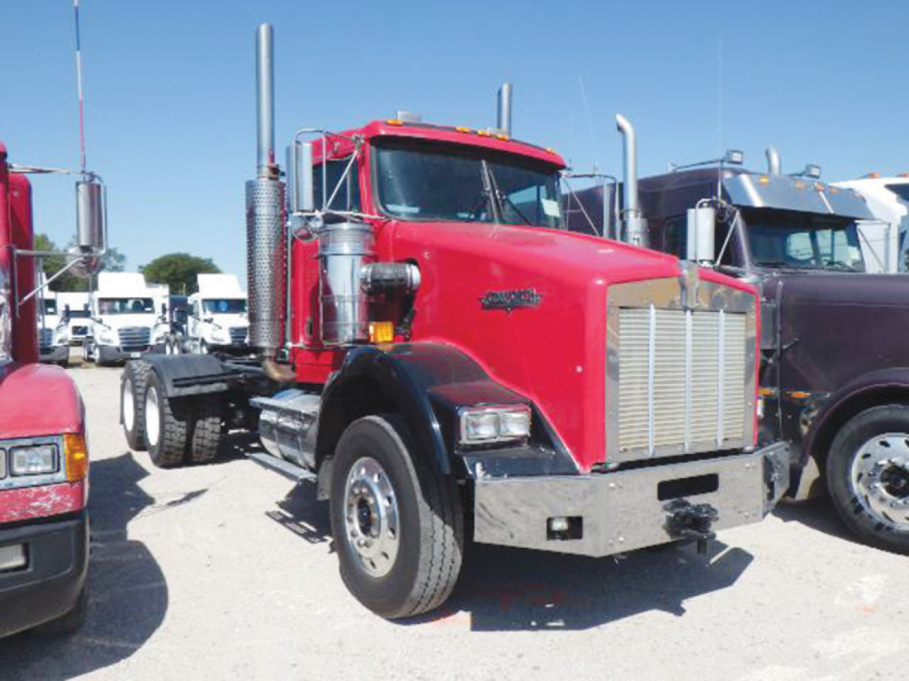 Kenworth T-800 conventional daycab tractor in red with chrome grille, dual vertical exhaust stacks, large front bumper, parked among other trucks.