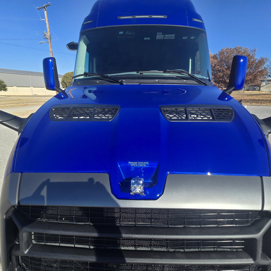 Front of a blue Mack PR64T76H truck showing hood vents, grille, and windshield; side mirrors visible.