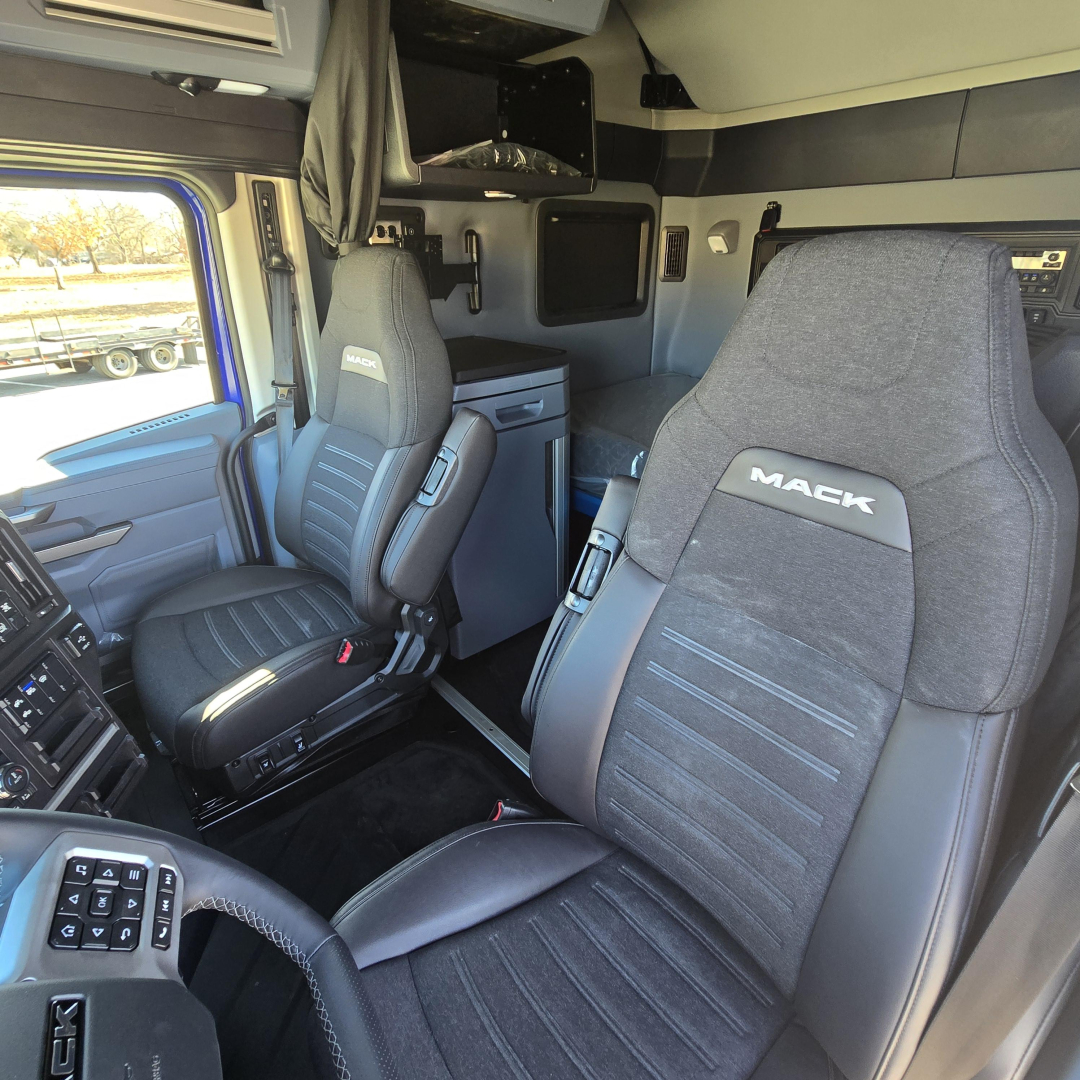 Mack PR64T76H truck interior showing driver and passenger seats with dashboard and storage cabinet behind, modern cab with dark upholstery.