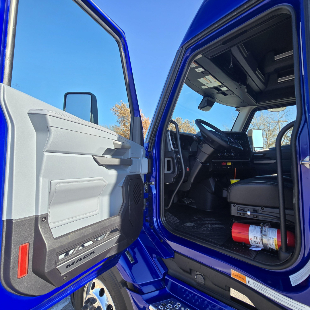 Truck cab interior visible through open door, steering wheel and dashboard, blue exterior. Door panel shows gray plastic with a small storage pocket and speaker. Floor mat and red fire extinguisher visible under dash.
